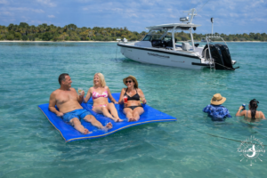 Guests relaxing on a floating water mat at a Sanibel Island sandbar while Florida Gulf Discovery's Axopar 29 is anchored nearby in clear Gulf waters.