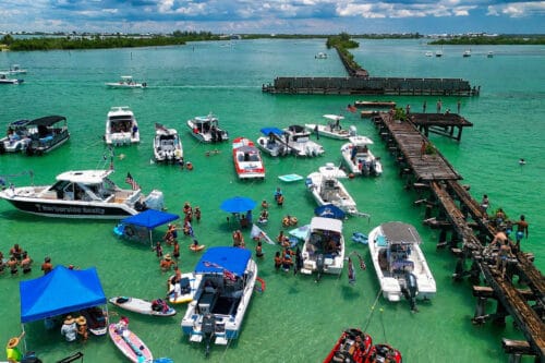 Drone view of boats anchored at Boca Grande Sandbar during the Independence Day celebration in Southwest Florida, with people enjoying the shallow turquoise waters near the historic railroad trestle.