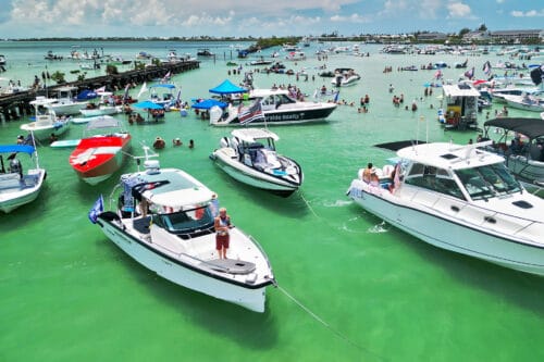 Drone view of dozens of boats anchored at Boca Grande Sandbar during the Independence Day celebration, with people swimming in the crystal-clear turquoise waters of Southwest Florida.