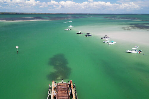 Aerial drone view of the Placida Trestle and Boca Grande Sandbar in Gasparilla Sound, Florida, with boats anchored in shallow emerald water.