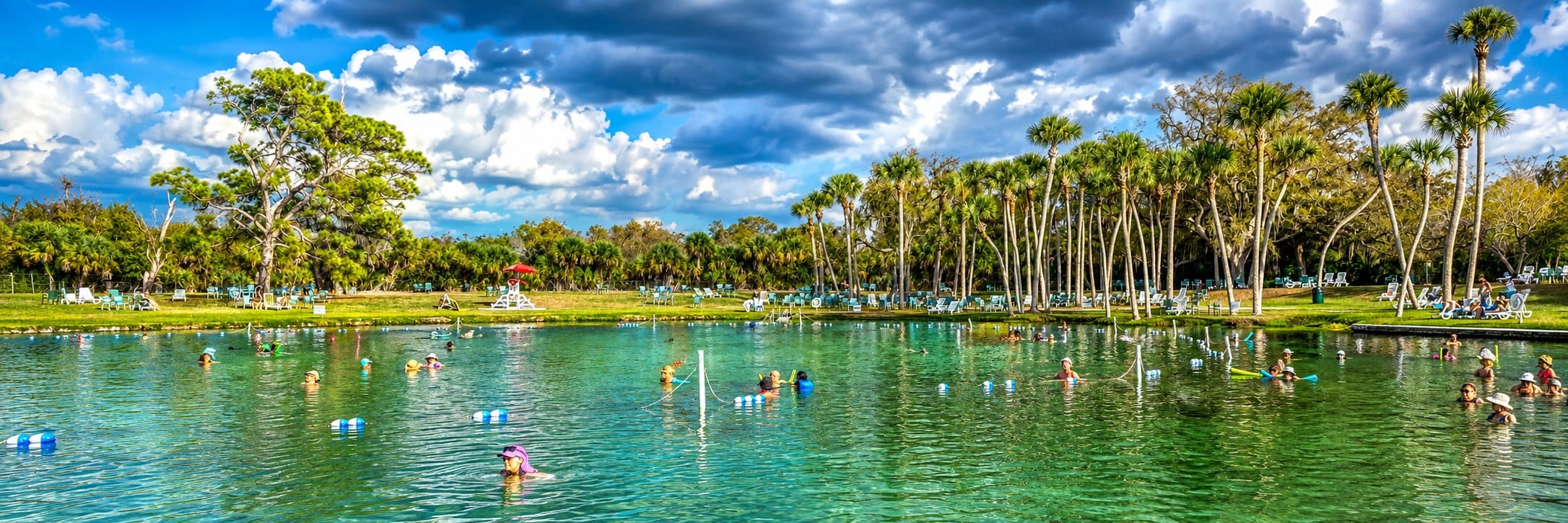 Panoramic view of Warm Mineral Springs in North Port, Florida, showing visitors enjoying the naturally warm mineral waters beneath dramatic summer storm clouds.